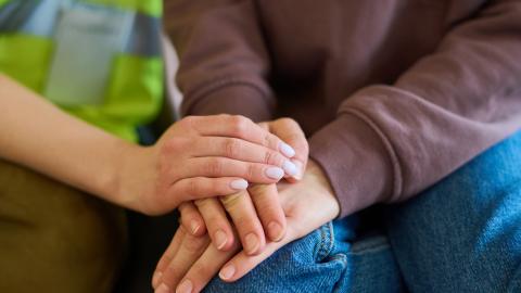 Hand of female volunteer in uniform covering that of young refugee in blue jeans and purple sweatshirt sitting next to her