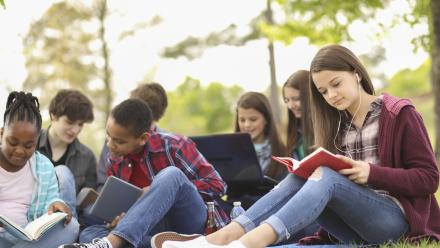Group of teenagers reading books while sitting on the ground outside.